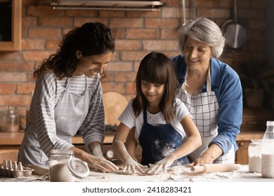 Hands in flour. Friendly intergenerational family of 3 diverse age females engaged in cooking short pastry biscuits. Young mom senior granny assist little girl stretch dough for pie on kitchen table - Powered by Shutterstock