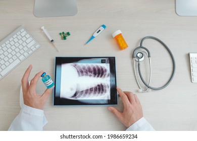 Hands Of General Practitioner Holding Vial Of Vaccine Against Coronavirus When Analyzing Chest X-ray Of Patient On Tablet Computer, View From Above