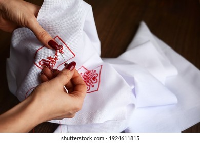 Hands Of A Girl,  Woman  Sew A Small Embroidery Pattern On A White Canvas. The Ornament Is Made With Red Floss Threads. Cross Stitch Type