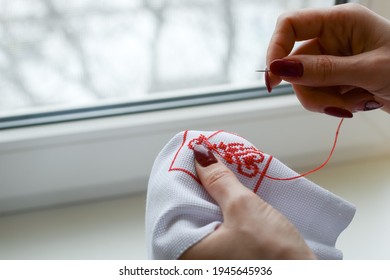 Hands Of A Girl, Woman Sew A Small Embroidery Pattern On A White Canvas. The Ornament Is Made With Red Floss Threads. Cross Stitch Type