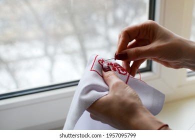 Hands Of A Girl, Woman Sew A Small Embroidery Pattern On A White Canvas. The Ornament Is Made With Red Floss Threads. Cross Stitch Type