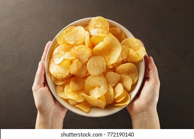 Hands giving bowl of potato chips on brown background. Concept of snack food. Top view.