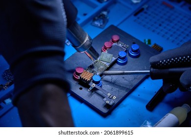 Hands In Gloves Of Cellphone Technology Engineer Work On Chipset Pins Of Removed Microprocessor From Smartphone Motherboard With Soldering Iron And Hot Air Heater. Macro Photo, Selective Focus