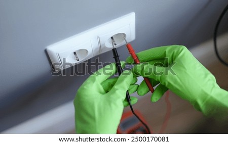 Hands in green gloves plug the voltmeter into an outlet, close-up. Home electrical wiring repair, electrician equipment