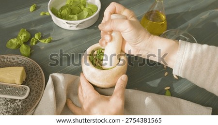 Hands grinding pine nuts and basil in marble mortar for Genovese pesto. Close-up of person using stone pestle to crush ingredients for traditional Italian sauce on wooden table. Making authentic pesto