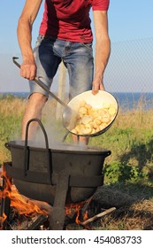 Hands Of Guy Pours A Ladle From Bowl Chopped Potatoes Into The Boiling Cauldron On The Fire In The Black Soot