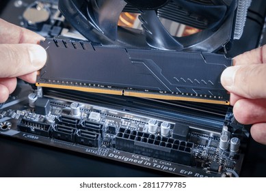 Hands hold a DDR memory module above a DIMM slot on a motherboard. SATA ports, an ATX power connector, and a CPU fan are visible. Modern PC assembly or upgrade