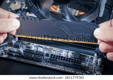 Hands hold a DDR memory module above a DIMM slot on a motherboard. SATA ports, an ATX power connector, and a CPU fan are visible. Modern PC assembly or upgrade