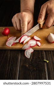 Hands hold a knife while cutting radishes on a wooden board. The kitchen table is dark, and pieces of radish are scattered around. Fresh vegetables are present.