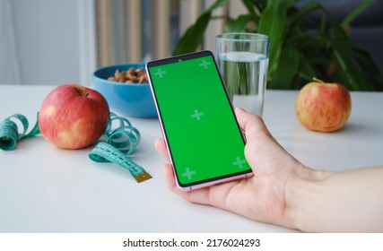 Hands hold a mobile phone with a green screen on the background of apples, dry breakfast and a glass of water. Clean eating and exercise for good health. Organic food, diet concept. - Powered by Shutterstock