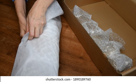 Hands Hold Packing Material And Wrap Glassware Into Soft Packing Due To Prepare For Moving Day. Glass Tableware Wrapped Into Plastic Bubble Wrapping Inside Cardboard Box