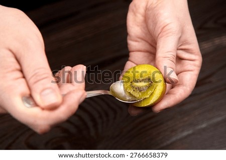 Hands hold a spoon while preparing to scoop out green kiwi fruit flesh from a halved kiwi against a dark wooden surface