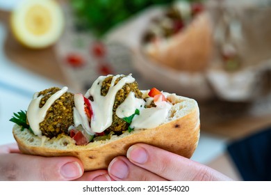 Hands Holding An Authentic Half Of Pita Bread Sandwich Filled With Fresh Falafel Balls, Chopped Salad And Drizzle Of Tahini Sauce On Top. Selective Focus On Chickpea Falafels In Fluffy Pita, Colorful.