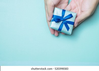 Hands Holding Blue Wrapped Gift Box With Blue Ribbon As A Present For Christmas, New Year, Mother Day Or Anniversary On Blue Table Background, Top View