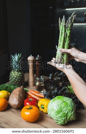 Hands are holding a bunch of asparagus while various vegetables and fruits are arranged on a wooden table. Items include lettuce, tomatoes, carrots, and more. The setting looks bright.