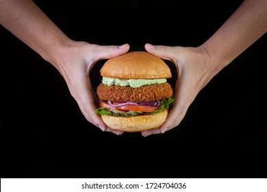 Hands holding a chicken burger with lettuce, tomato, purpple onion and handmade mayo on black backgorund. Delicious.