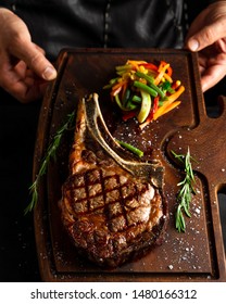 Hands Holding Cooked Tomahawk (long Bone Ribeye) Steak On A Serving Board. Low Key Image, Vertical Orientation