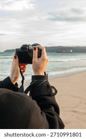 Hands holding a digital camera while photographing seascape on sandy beach, capturing outdoor moments with modern photography equipment and natural coastal scenery.
