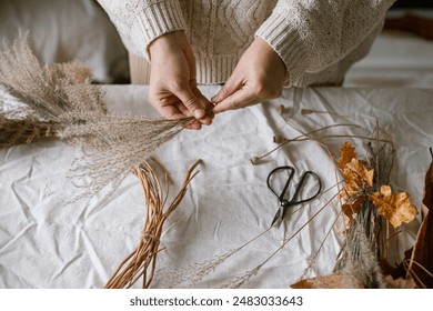 Hands holding dried grass, herbs, leaves, ribbon and scissors on rustic table. Making stylish autumn wreath. Fall decor and arrangement in home. Thanksgiving and Halloween - Powered by Shutterstock