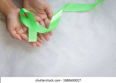 Hands Holding Emerald Green Or Jade Green Ribbon On White Fabric Background With Copy Space, Symbol For Liver Cancer Awareness, World Cancer Day. Healthcare Or Hospital And Insurance Concept.