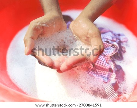 Hands holding foam bubbles while washing clothes in a red basin