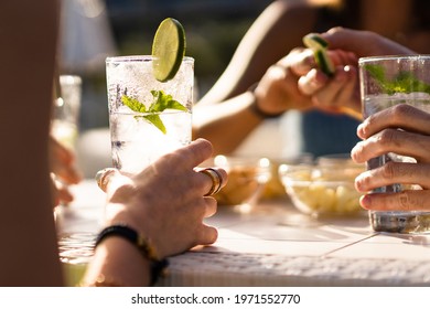 Hands Holding A Fresh Alcoholic Long Drink Cocktail On A Table. Gathering Of Friends In An Outdoor Pub In The Summer.