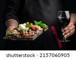 Hands holding a glass of wine and a wooden board with different kinds of cheese and ham, prosciutto, jamon salami, Antipasto Dinner or aperitivo party.