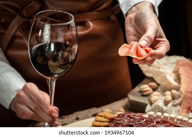 Hands Holding A Glass Of Wine And A Wooden Board With Different Kinds Of Cheese And Ham, Prosciutto, Jamon Salami, Antipasto Dinner Or Aperitivo Party Concept.