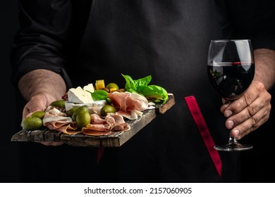 Hands holding a glass of wine and a wooden board with different kinds of cheese and ham, prosciutto, jamon salami, Antipasto Dinner or aperitivo party. - Shutterstock ID 2157060905