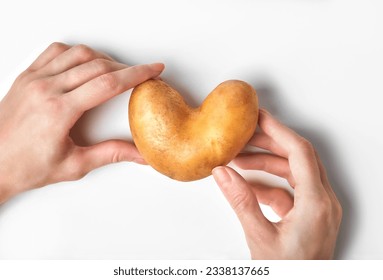 Hands holding heart-shaped potato above white background. Ugly food, funny vegetable concept. Horizontal orientation, top shot.