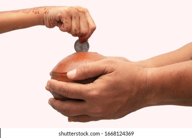 Hands Holding Indian Penny Bank Made With Mud Clay And Child Hand Adding Coin Into The Earthen Pot Or Terracotta Piggy Bank On Isolated Background - Concept Showing Of Savings, Investment Or Donating