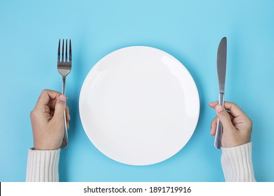 Hands holding knife and fork above white plate on blue background., dieting, weight loss, dining and kitchenware concept