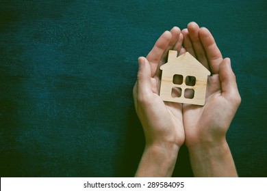 Hands Holding Little Wooden House On Blackboard Background. Symbol Of Construction, Security Or Sweet Home Concept