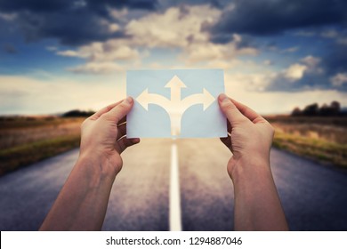 Hands holding paper with arrows crossroad symbol splitted in three different directions. Choose the correct way between left, right and front. Difficult decision concept, over asphalt road background.