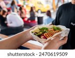 Hands holding a paper container filled with Asian street food, featuring noodles and vegetables, against a blurred background of a busy market