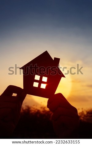 Hands holding paper house on sunset sky, family home, homeless housing, and home protection insurance concept, international day of families, new life, foster home care, and real estate.