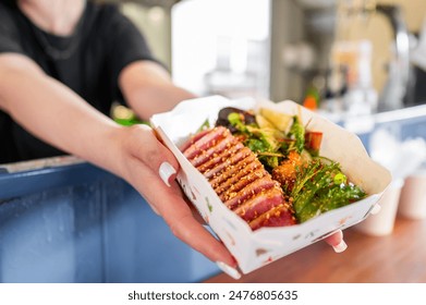 Hands holding a paper tray with sliced grilled tuna and green garnish, suggesting a casual dining or street food scene - Powered by Shutterstock