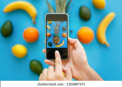 Hands Holding Phone And Taking Photo Of Pineapple In Sunglasses And Bananas, Oranges And Avocado On Blue Paper, Trendy Flat Lay. Stylish Food Photography. Instagram Fruit Photo