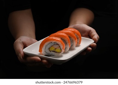 Hands holding plate of Uramaki sushi or California salmon roll on black background. Japanese food style concept - Powered by Shutterstock