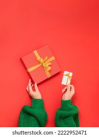 hands holding red gift box with golden ribbon bow and white plastic card on red background. Flat lie, copy space