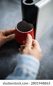 Hands holding a red mug while brewing hot espresso coffee from a modern coffee machine. Morning routine for a caffeine dose