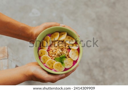 Hands holding round plate of granola with bananas and berries on light concrete background. The concept of a healthy and delicious breakfast. Smoothie bowl in hands flatlay