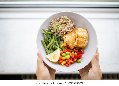 Hands Holding Salmon And Buckwheat Dish With Green Beans, Broad Beans, And Tomato Slices. Nutritious Dish With Vegetables And Fish From Above. Healthy Balanced Diet