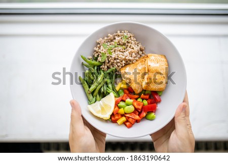 Hands holding salmon and buckwheat dish with green beans, broad beans, and tomato slices. Nutritious dish with vegetables and fish from above. Healthy balanced diet