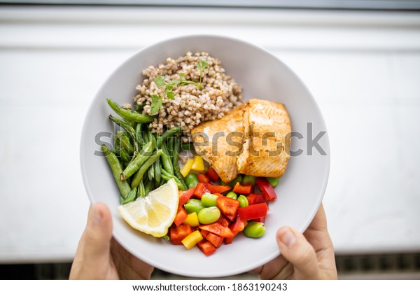 Hands holding salmon and\
buckwheat dish with green beans, broad beans, and tomato slices.\
Nutritious dish with vegetables and fish from above. Healthy\
balanced diet