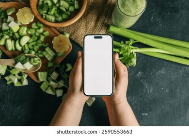Hands holding a smartphone with a blank screen over a table with fresh green vegetables, including celery, cucumber, and apple. A green smoothie in a glass and a cutting board  - Powered by Shutterstock - Get 15% off with code: PIKWIZARD15