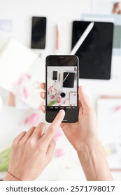 Hands holding a smartphone, capturing a flat lay of tech gadgets and stationery. Focus on phone, tech, and stationery. Creative workspace with tech and stationery. Content creator taking photo of desk