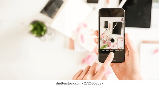 Hands holding a smartphone, capturing a flat lay of office supplies. Smartphone photography of notebooks, coffee, and flowers. Creative flat lay photography. Content creator taking photo of desk.