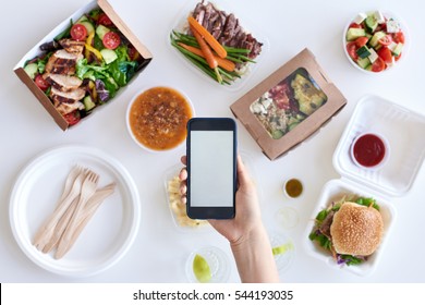 Hands holding smartphone mobile cellphone over different types of gourmet takeout, food delivery order app application concept