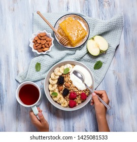 Hands Holding Spoon Of Healthy Breakfast With Granola, Berries And Yogurt And A Cup Of Tea, Top View, Overhead, Healthy Eating, Morning Snack